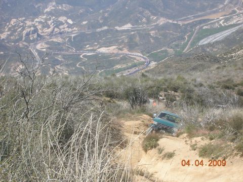 1985 Ford Bronco - A green pickup truck navigates a steep dirt path, with a winding road and valley visible beyond.