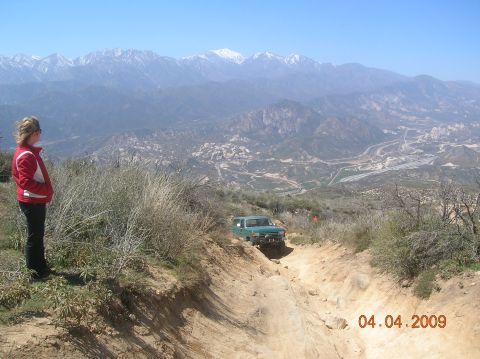 1985 Ford Bronco - A person watches a green SUV navigate a challenging off-road trail with a vast mountain landscape behind.