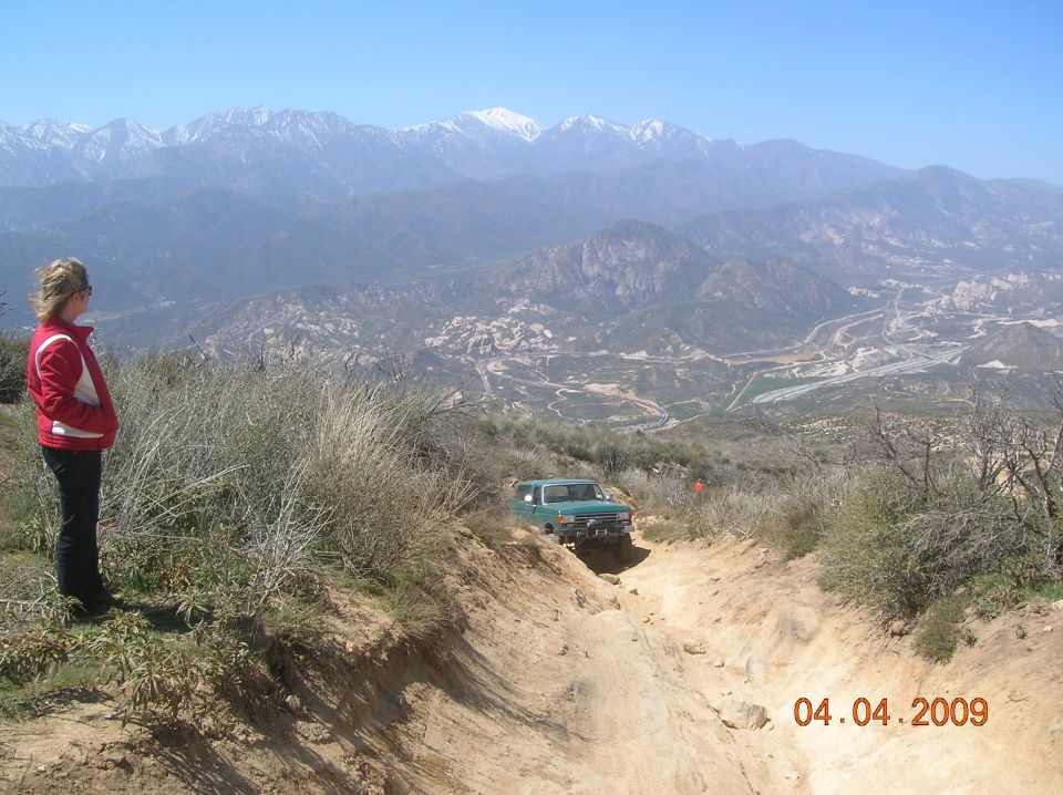 Green SUV driving down a steep dirt trail with mountains in the background. Woman stands on the side of the trail