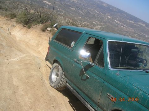 1985 Ford Bronco - A green SUV navigates a steep, dusty off-road trail with a scenic mountain vista.
