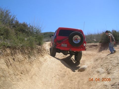 1985 Ford Bronco - A red SUV navigates a steep dirt trail with a spare tire mounted on the back.