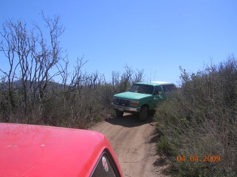 1985 Ford Bronco - A green 1990s Ford Bronco navigates a dusty trail surrounded by dry brush and trees.