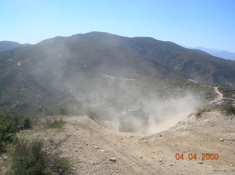1985 Ford Bronco - An off-road vehicle navigates a dusty mountain trail, creating a large dust cloud behind it.