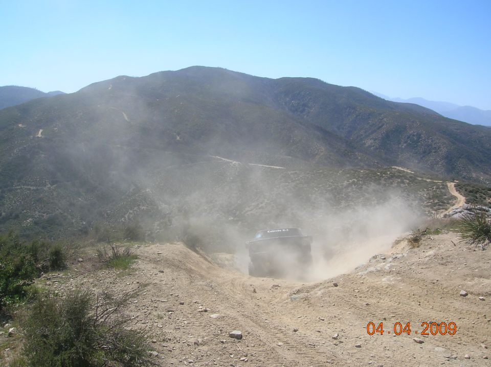 SUV driving on dusty dirt road in mountains, kicking up a lot of dust. Date stamp reads 04.04.2009