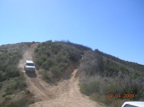 1985 Ford Bronco - Two vehicles navigate a steep, dusty off-road trail under a clear blue sky.