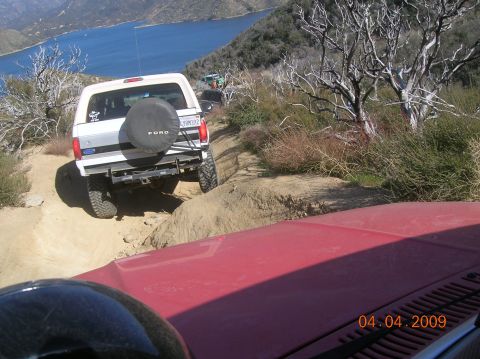 1985 Ford Bronco - White Ford Bronco navigating a steep dirt trail with a lake in the background. Another vehicle is ahead on the path.