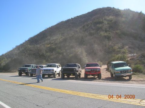 1985 Ford Bronco - A group of SUVs are parked on the side of a road with a dusty hill in the background.