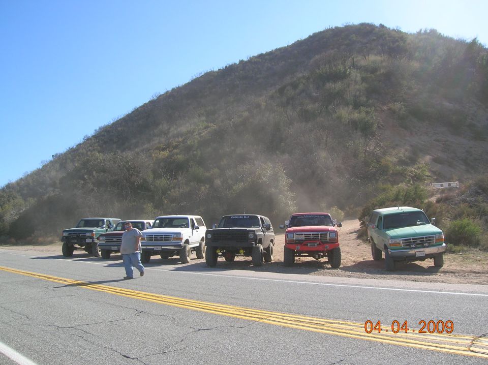 several SUVs parked on the side of a road with a dusty hill behind them. A person walks across the road in the foreground