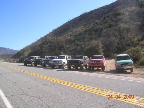 1985 Ford Bronco - A line of SUVs, including several Ford Broncos, are parked on the side of a road on a sunny day.