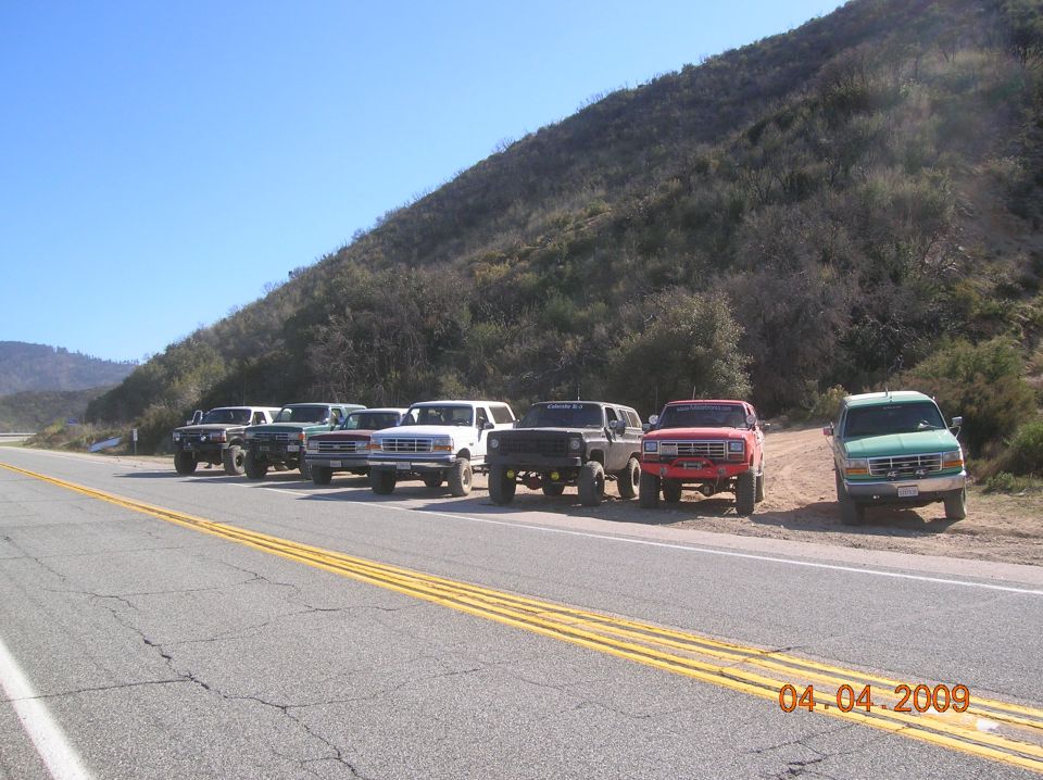 Seven SUVs parked on a dirt shoulder next to a highway, with a grassy hill in the background