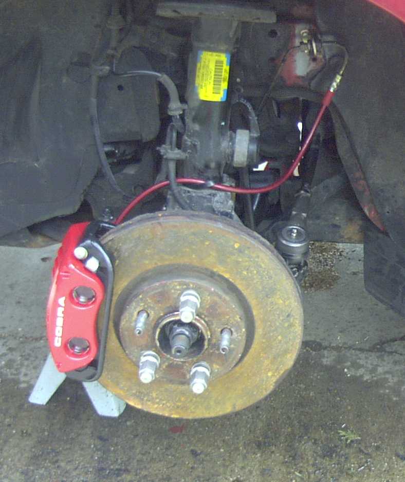 Red brake caliper labeled COBRA, mounted on a rusty brake rotor. The suspension and brake lines are visible