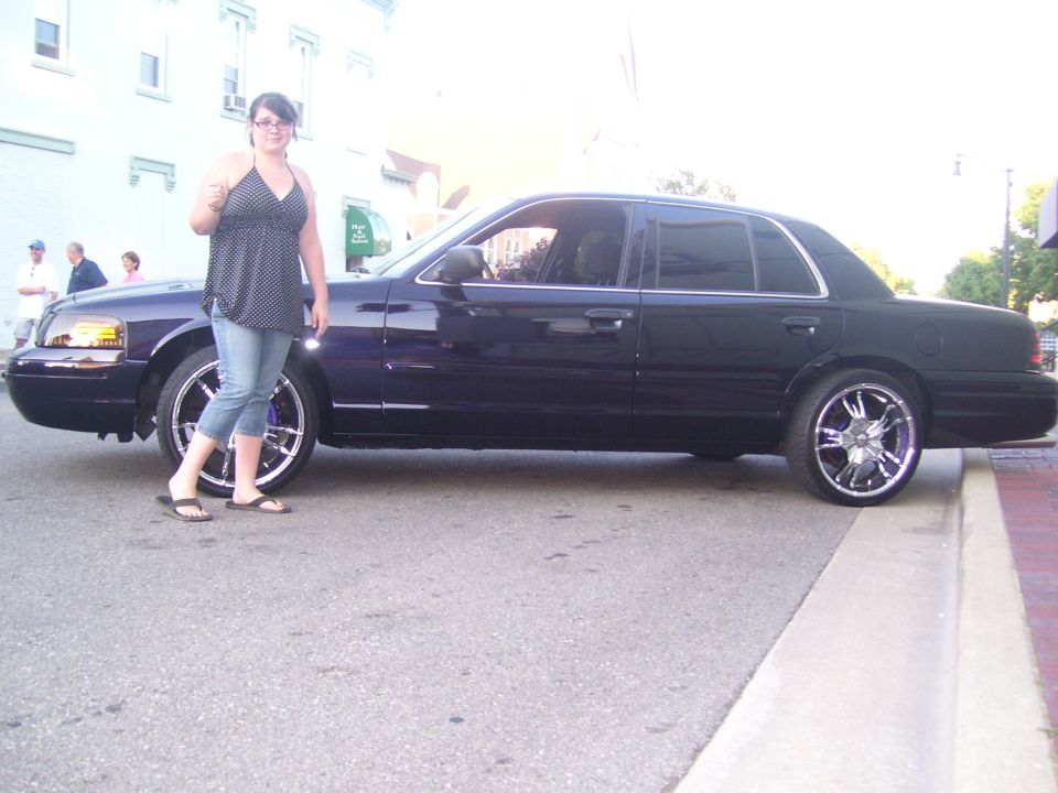 Dark Ford sedan in side profile with chrome wheels, woman posing beside it on a street.