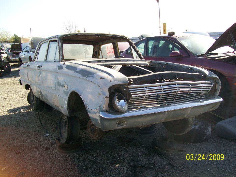 1960 Ford Falcon sedan, front view, missing hood and engine, in junkyard. Another car is parked behind it