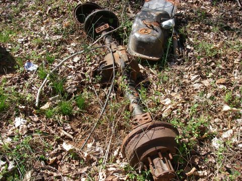 1978 Ford Bronco - Discarded vehicle parts, including an axle and fuel tank, are scattered on the ground amidst foliage.