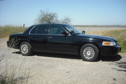 2003 Ford Crown Victoria - A black 2003 Ford Crown Victoria sedan is parked on a gravel road with a tree and field in the background.