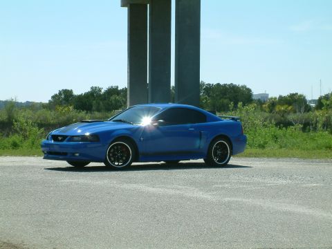 1992 Ford Bronco - A blue Ford Mustang coupe is parked on a paved road next to a concrete structure. The car has black racing stripes on the hood and black ri…