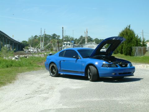 1992 Ford Bronco - A bright blue Ford Mustang coupe with its hood open is parked on a gravel lot. In the background, a highway overpass and several boats are…