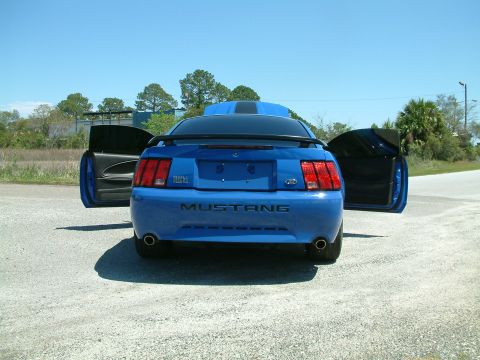 1992 Ford Bronco - The rear view of a bright blue Ford Mustang Mach 1 with its doors open, parked on a gravel surface under a clear blue sky. The car has a sp…