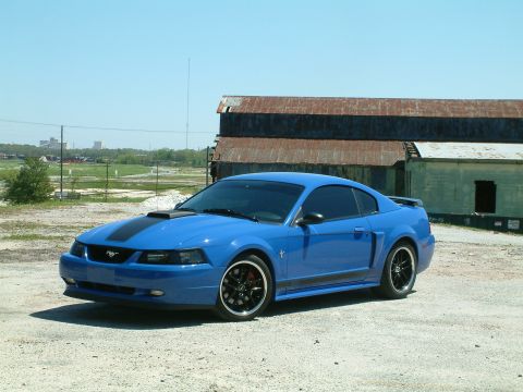 1992 Ford Bronco - A bright blue Ford Mustang coupe is parked on a gravel lot in front of a weathered, rusty industrial building. The car features black racin…