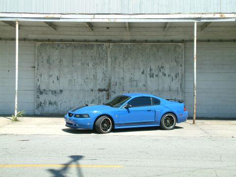 1992 Ford Bronco - A bright blue Ford Mustang coupe is parked in front of a large, weathered industrial garage door. The car has black racing stripes on the h…