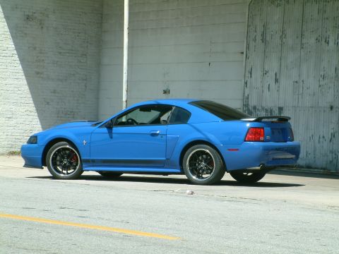 1992 Ford Bronco - A bright blue Ford Mustang is parked on the side of a road, with a corrugated metal wall behind it. The car has black racing stripes on its…