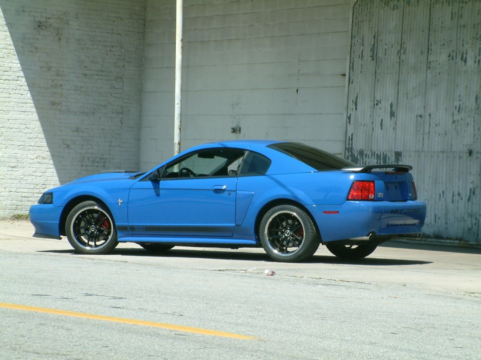 Blue Ford Mustang with black racing stripes and rims parked next to a corrugated metal wall.