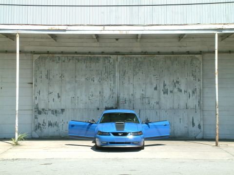 1992 Ford Bronco - A blue Ford Mustang with its doors open sits in front of a large, weathered corrugated metal garage door.