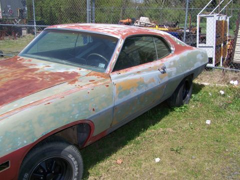 1978 Ford Bronco - A rusty, weathered coupe with significant paint deterioration sits parked on a grassy area next to a chain-link fence. The car's body shows…