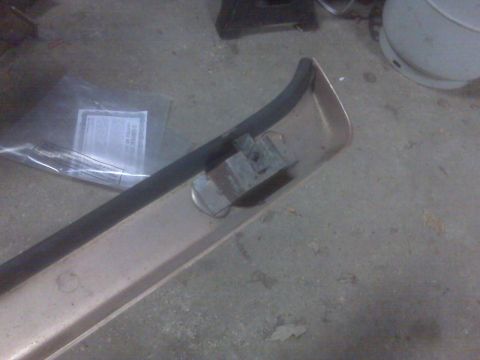 1995 Ford Bronco - A close-up view of a tan-colored bumper trim piece with a black rubber strip attached. A small metal bracket is visible on the trim.