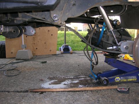 1996 Ford Bronco - The underside of a vehicle is shown, with suspension components, a differential, and a blue hydraulic jack supporting the frame. A wooden b…