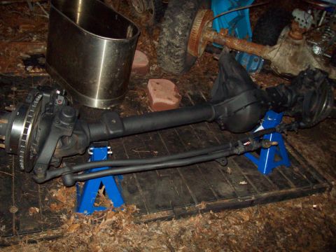 1989 Ford F-150 - A black front axle assembly with disc brakes and steering linkage is supported by two blue jack stands on a wooden surface covered in leave…
