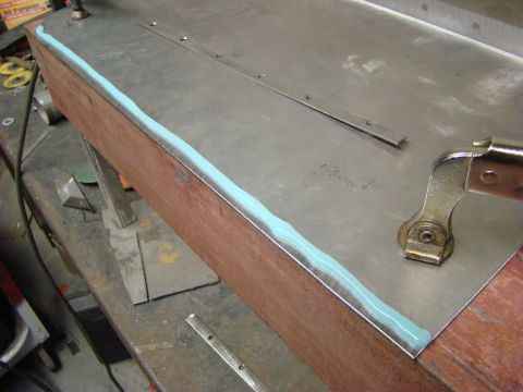 1948 Jeep Wrangler - A close-up view shows a wooden surface with a metal sheet attached, sealed with a wavy line of light blue sealant. A metal clamp is visible…