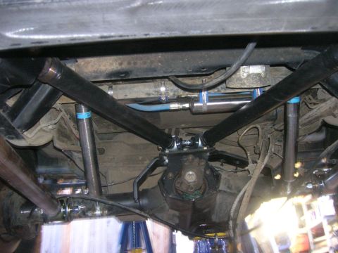 1996 Ford Bronco - Underside view looking up at a solid front axle with a four-link/radius-arm setup: black tubular links converge on a machined third-member…