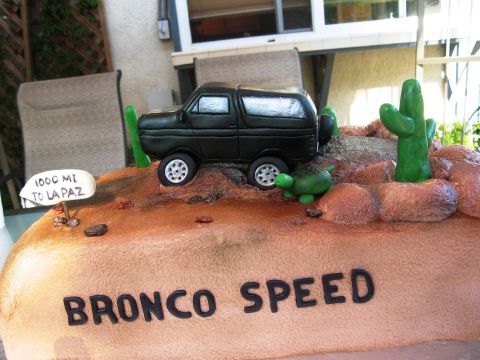1986 Ford Bronco - A novelty cake sculpted to look like a desert scene, featuring a black SUV-shaped fondant vehicle climbing rocky terrain with green cactus…
