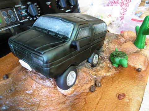 1986 Ford Bronco - A fondant cake sculpted as a black 1980s-style Ford Bronco with a lightbar on the roof, sitting on a desert-scene cake board with a green c…