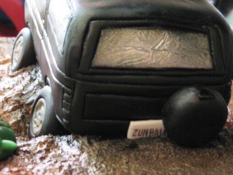 1986 Ford Bronco - Close-up of a black novelty cake shaped like a boxy SUV, with a molded rear window, wheels, and a small white tag reading 'ZUNBAJA' resting…
