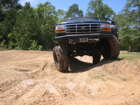 1995 Ford Bronco - A lifted, dark-colored 1995 Ford Bronco is shown from a low angle, driving on a dirt track with large tires and suspension visible. The veh…