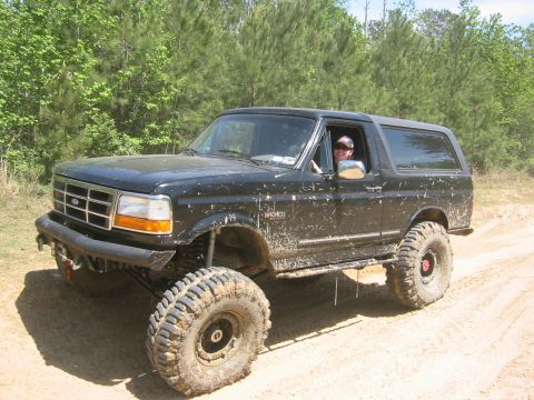 1995 Ford Bronco - A black, lifted Ford Bronco is shown driving on a dirt road with trees in the background. The vehicle is covered in mud, indicating off-roa…