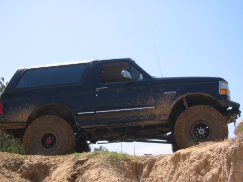 1995 Ford Bronco - A black 1995 Ford Bronco is shown in profile, driving off-road on a dirt hill with a clear blue sky in the background. The vehicle is cover…