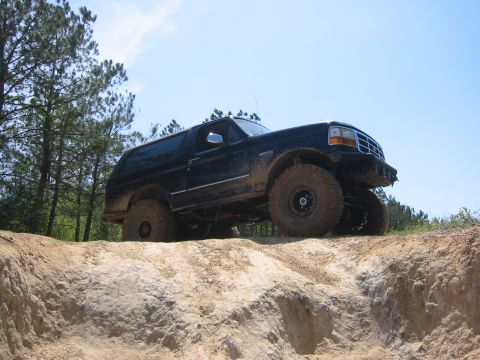 1995 Ford Bronco - A dark-colored Ford Bronco is shown from a low angle, driving up a steep dirt embankment with large off-road tires. The sky is clear blue w…