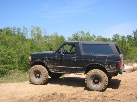 1995 Ford Bronco - A muddy black Ford Bronco with off-road modifications sits on a dirt path under a clear blue sky.