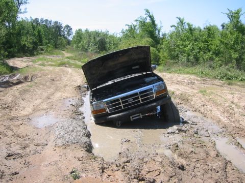 1995 Ford Bronco - This Ford Bronco is deeply stuck in a muddy rut on an unpaved road, with its hood open.