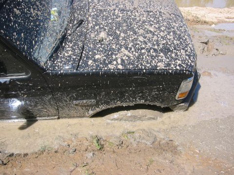 1995 Ford Bronco - A black 1995 Ford Bronco is deeply stuck in mud, with its tires submerged and the body splattered with dirt.