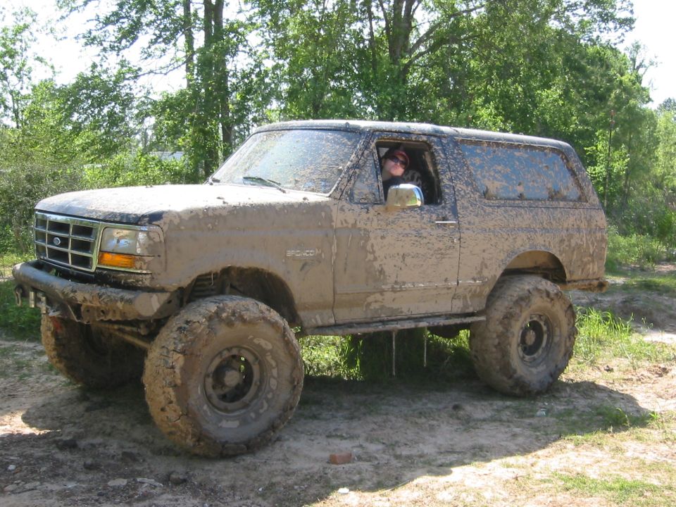 Mud-covered 1995 Ford Bronco parked on dirt with trees in background. Driver visible