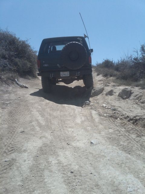 1982 Ford Bronco - A dark-colored Ford Bronco drives up a dusty, off-road trail under a clear blue sky.
