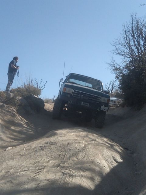 1982 Ford Bronco - A person watches an off-road vehicle navigate a steep, sandy trail under a clear blue sky.