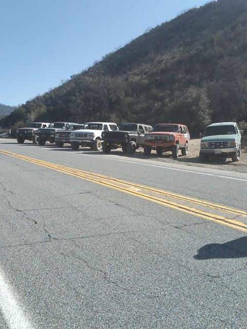 1982 Ford Bronco - A group of Ford Broncos and F-Series trucks are parked along a highway with a hillside in the background.