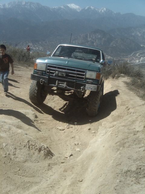 1982 Ford Bronco - A green pickup truck navigates a steep dirt trail with mountains in the background.