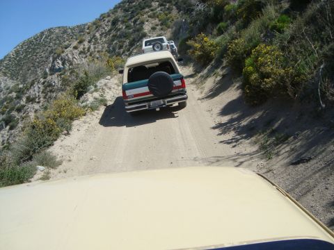 1986 Ford Bronco - A convoy of off-road vehicles navigates a narrow, winding dirt path on a mountainside under a clear blue sky.