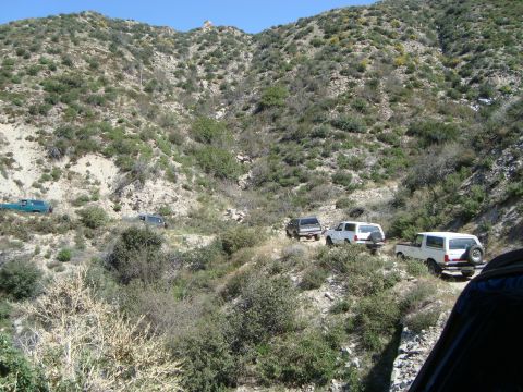 1986 Ford Bronco - Several SUVs navigate a dusty mountain trail under a clear blue sky.